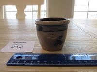 Front view of small ceramic crock on wooden surface with ruler for scale showing about 3 inches tall, and a maker's stamp.