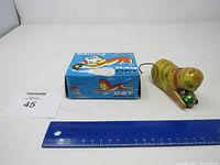 Entire lot showing the tin toy, original box, price card, and a ruler for scale.