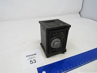 Full view of the dark metal coin bank resembling a small safe sitting next to a ruler for size reference