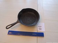 Top view of the entire cast iron skillet placed on a light wood surface with a blue ruler next to it for size reference, and a lot number card.