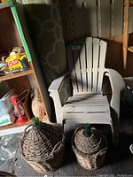 White Adirondack chair with outdoor rug rolled behind it and two wicker-covered demijohn bottles in front.
