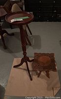 Two wooden accent pieces including a round tripod plant stand with raised edge and a small carved wooden stool with floral pattern.
