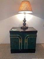 Nightstand with green panels, black top and base, gold trim, and a turned wooden lamp with fabric shade on top, lit.