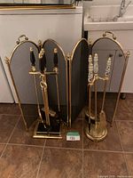 Front view of two brass three-panel fireplace screens and two fireplace tool sets with different handle styles.
