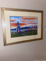Framed art featuring a colorful print of an Inukshuk sculpture with city skyline and sunset sky, mounted with a cream mat and wooden frame.