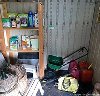 Photo shows a corner of a shed with wooden shelving stocked with various garden chemicals, a green broadcast spreader, two red gasoline cans, and a yellow electric pressure washer on the floor.