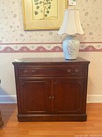 Full view of the wooden cabinet with ceramic floral lamp on top in a room with floral wallpaper and wooden floor.