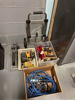 Overview photo of lot including dolly, manual tile cutter base, crates with tools, and box with cables