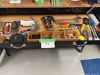 Table showing hand staplers, pliers, fasteners, utility knives, measuring tape, and assorted hardware.
