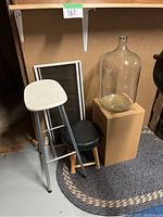 Two stools: one white metal seat with metal legs, one black padded seat with wooden legs, next to two large clear glass bottles on a cardboard box.