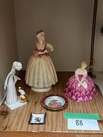 Overview of five bone china items on wooden shelf: two female figurines, tall white figurine with gold bird, two small decorated plates