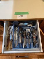 Open kitchen drawer with a blue Rubbermaid cutlery tray filled with assorted cutlery: forks, spoons, various knives with wooden and metal handles.