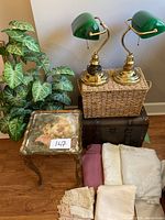 Photo showing pair of vintage green bankers desk lamps with pull chains on top of a wicker basket, decorative small painted wooden table with classical scene, dark vintage suitcase, and several folded large tablecloths/fabrics in light colors.
