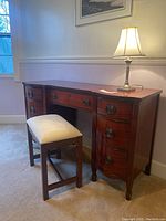 Front view of the vintage wooden pedestal writing desk with upholstered stool and table lamp on top.