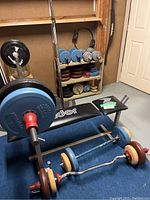 Workout bench with barbells and weight plates on floor and shelf unit with weights in background