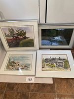Four framed prints shown leaning against cabinetry. Includes colorful landscape with trees, river through forest, coastal lighthouse, and house architectural renderings.