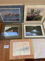 Five framed prints showing a mix of landscapes including city skyline, rural house, lake, river, and a birch tree winter scene, grouped on floor.