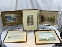 Five framed watercolor paintings and prints arranged in a group, sitting on table with white backdrop.