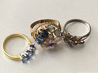 Photo showing three rings side by side on a flat surface: one gold-tone ring with blue rectangular glass stones, one gold-tone ornate ring with multicolored glass stones, and one sterling silver ring with floral design.