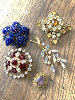 Top view showing front of five costume jewellery brooches on wooden surface: blue Austria stamped, amethyst glass with gold lace border, circular red and clear rhinestone, amber colored diamond shaped, wheat motif brooch.