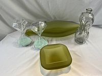 Full set of glass home decor items: two sand timers, decanter, two olive green serving pieces photographed on white surface