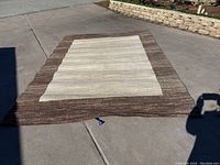 Full view of the large area rug laid flat outside, showing the beige center and brown border with scalloped edge design on the border.
