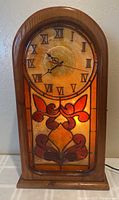Front view of the clock showing the wooden frame, Roman numeral dial, and stained glass illuminated by the backlight.