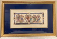 Full view of framed embroidered angel needlepoint artwork with gold frame and blue matting on a table.