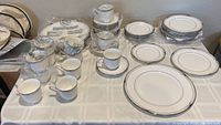 Wide view of 49 pieces of Lenox Erin pattern bone china including plates, cups and saucers on white cloth surface, some still partly wrapped in plastic and bubble wrap.