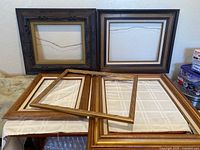Five vintage picture frames arranged on a surface, two dark carved wood frames in back and three wooden frames laid in front without glass.