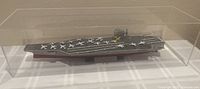 Side view of USS John C. Stennis aircraft carrier scale model with multiple miniature aircraft on deck under clear plexiglass cover on a white surface.