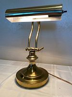 Front angle view of vintage brass desk lamp on white table, showing cylindrical shade, curved supports, and round base.