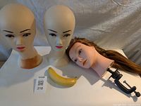 Three mannequin heads on a table including two without hair and one with long hair and a clamp. A banana is placed for size reference.
