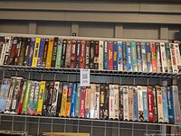 Photo of two shelves filled with 70 VHS tapes showing spines with various movie titles, condition good used.
