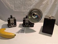 Three cameras against neutral background showing entire lot with a banana for scale. Two Brownie Hawkeye cameras, one with large flash, and one antique Brownie box camera.
