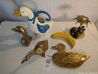 Five duck-themed decorative items: 4 brass and 1 ceramic, including a ceramic duck pitcher with blue details, brass duck figurine, brass duck tray, brass duck head bookend, and a brass trivet with grapes and umbrella.