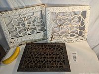 Three cast iron heat registers placed upright with a banana for scale showing size. Two wall-mounted white ornate decorative registers behind one flat black floor register.