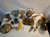 Photo showing the full array of brass items from a top-down angle with banana for scale, including boot planter, brass kettle, goblets on tray, pan, incense burners, pitcher, cup, mortar, and roasting fork.