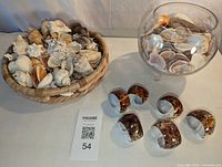 Wicker basket containing assorted shells, footed crystal bowl with shells, and six cut shell napkin holders arranged together on a white surface with a banana for scale.