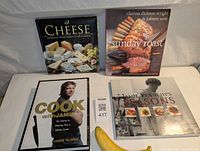 Four cookbooks displayed together with a banana placed in front for scale. Titles visible include 'Cook with Jamie' by Jamie Oliver, 'Cheese', 'Sunday Roast', and 'Seasons'.