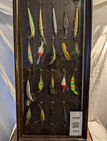 Front view of all 19 fishing lures displayed hanging inside a wooden and glass case, showing different styles and colors.
