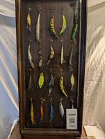 Front view of 20 fishing lures in a frame, showing diverse colors, sizes, and shapes hung on hooks with treble hooks attached.