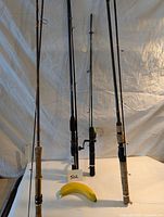Four fishing rods standing upright with varying handle styles and reel seats, banana in foreground for scale.