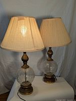 Pair of matching lamps with pleated beige shades, clear crackled glass spherical bodies, and ornate brass metal bases shown side by side.