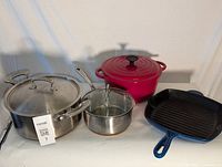 Photo showing set of two stainless steel pots with lids, red cast iron Dutch oven, and blue cast iron grill pan viewed from the front.