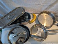 Photo showing the large stainless steel sautee pan with lid, smaller stainless steel pans, and an elongated fish poacher with lid and vented steam rack. Banana included for scale, not for sale.