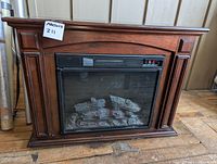 Full frontal photo showing the electric fireplace with wood-finish cabinet and glass panel with fake logs.