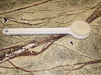 Photo of a white 14-inch back scrubber with a long handle and rounded soft bristle brush head placed on a marbled surface.