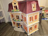Exterior front and side view of the Playmobil Large Grand Mansion showing detailed yellow and white facade, red shingled roof, balconies with flowers, orange shutters, windows and front steps.