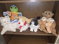 Shelf of assorted battery-operated stuffed animals including white fluffy cat, white dog with red bow, black and white cat, tan donkey, two brown baby monkeys, beige dog, and LeapFrog Learning Friend Todd frog plush in orange outfit.